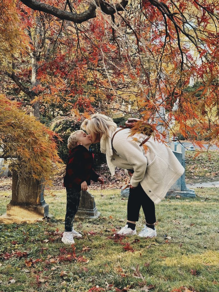 A woman kissing a young child beneath orange-red maple branches on an autumn day.