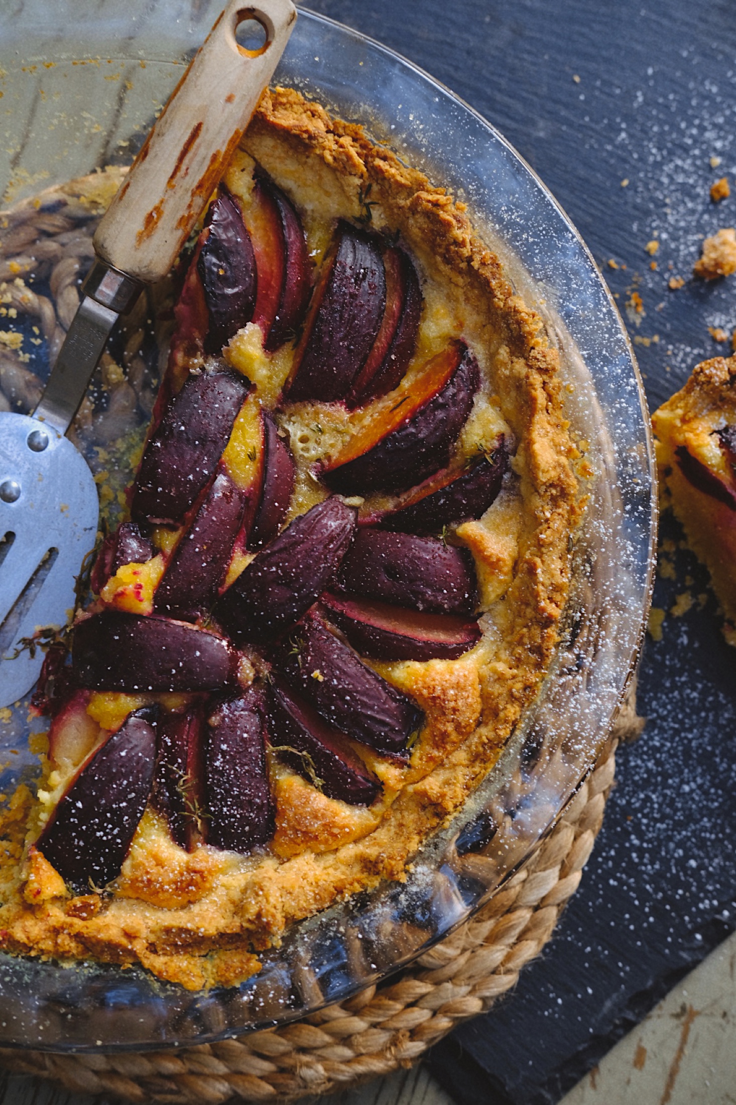 Overhead view of a plum and thyme frangipane tart in a glass pie dish, with deep purple fruit arranged over golden almond cream.
