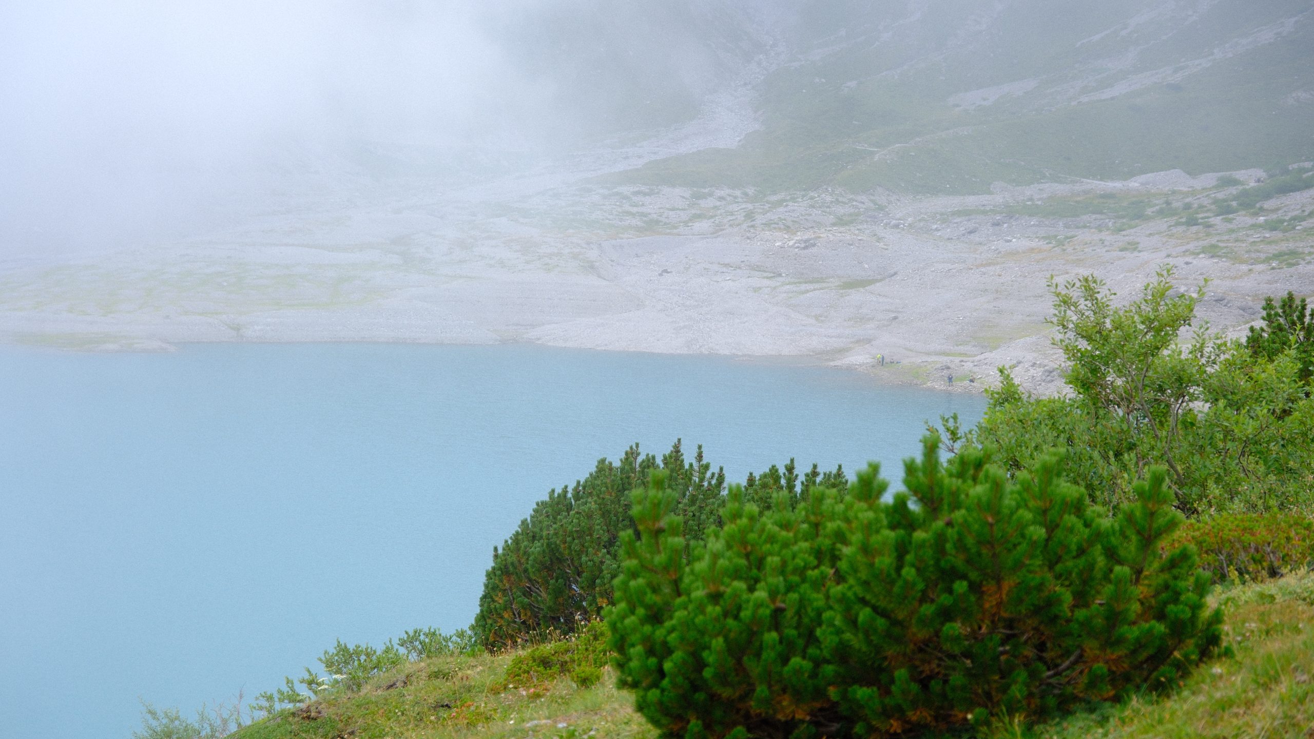 A pale rocky shore curving around turquoise Lünersee beneath low cloud.