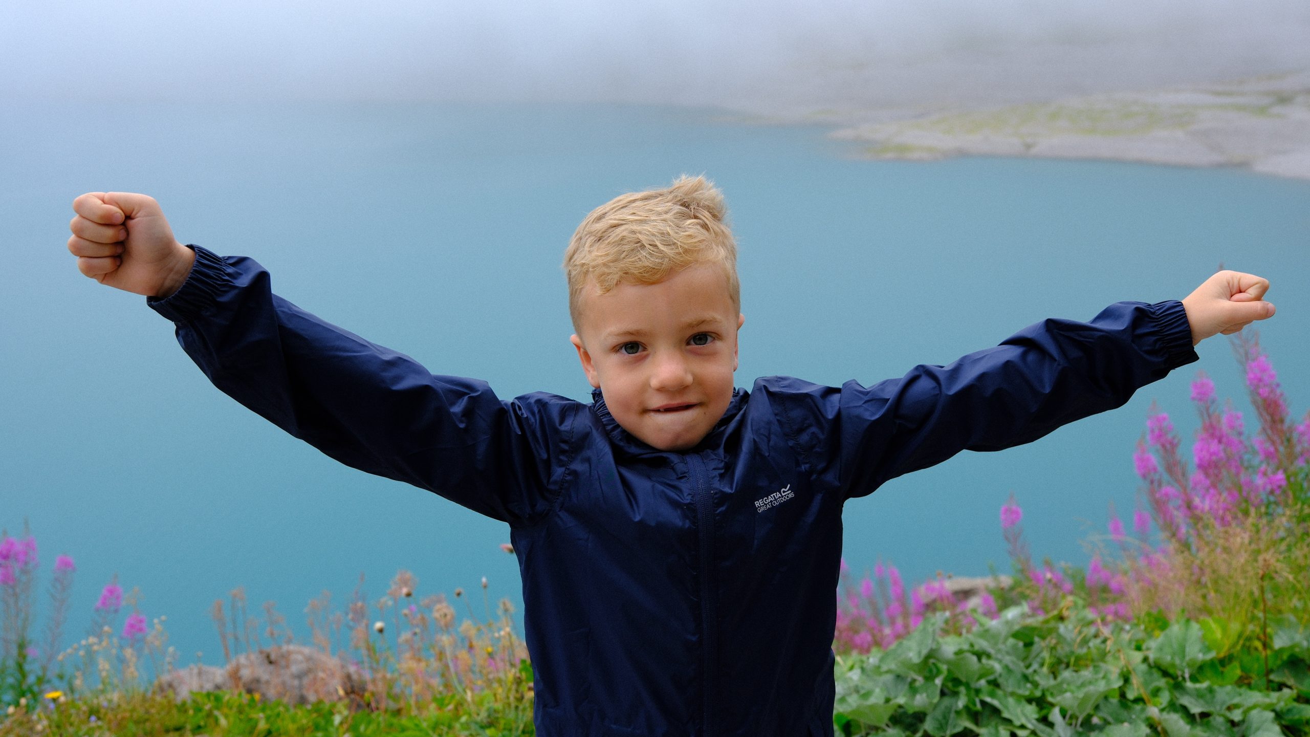 Chris standing with his arms raised above turquoise Lünersee and pink fireweed.
