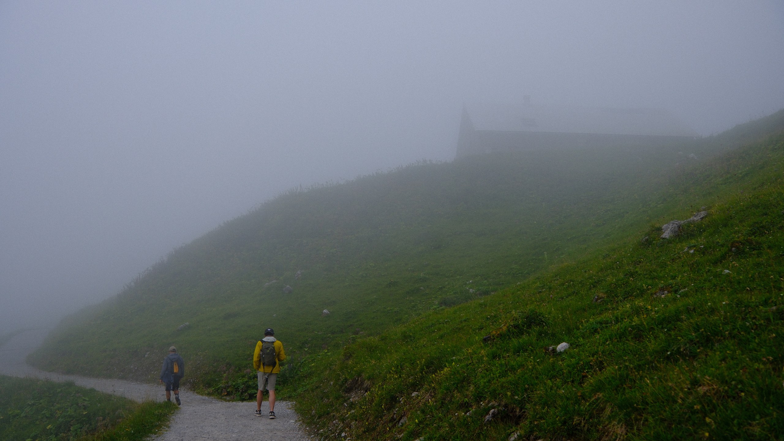 Two walkers in bright jackets disappearing along a mountain path into dense mist.