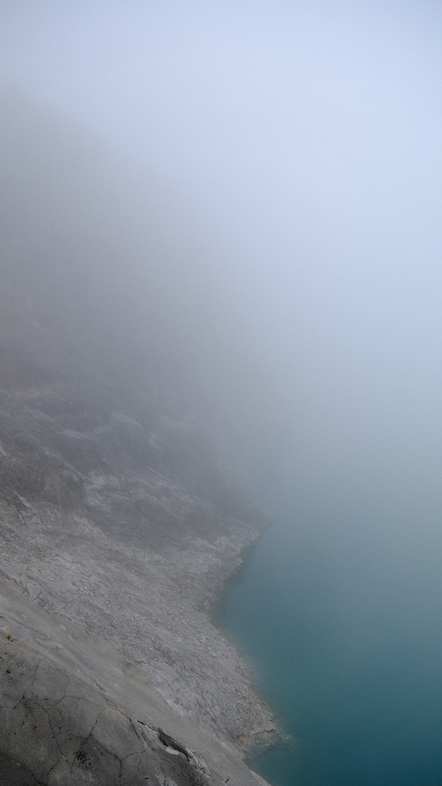 A vertical glimpse of turquoise Lünersee beneath a steep rock wall and heavy mist.