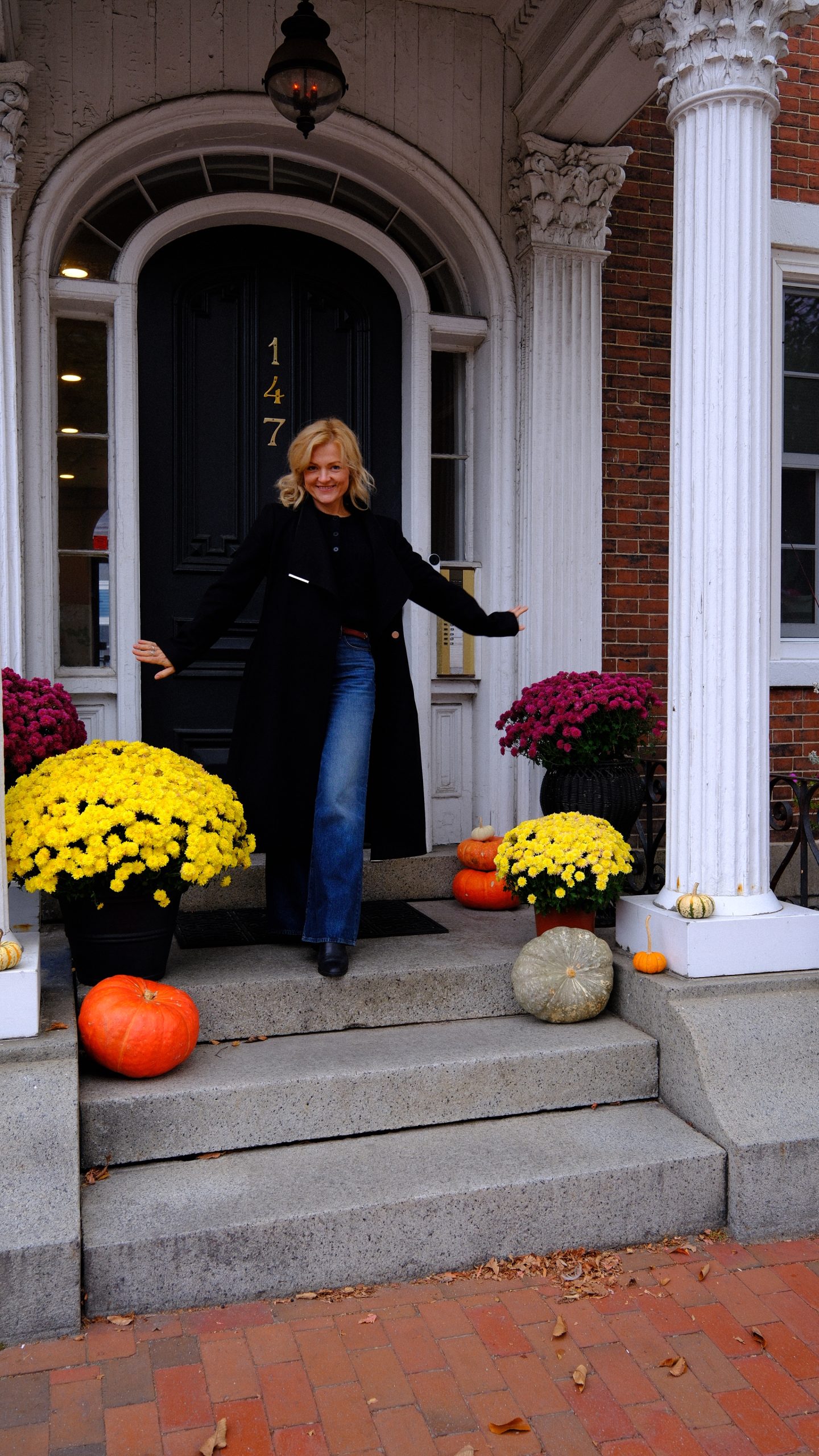 Karolina standing on an autumn porch surrounded by pumpkins and chrysanthemums